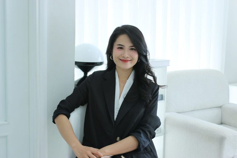 Vũ Hà Trang mặc blazer đen ngồi tựa bên cửa sổ rèm trắng