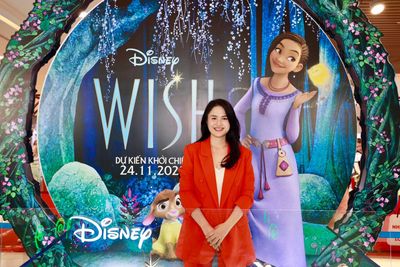 Vũ Hà Trang áo vest cam đỏ đứng trước standee phim Wish của Disney