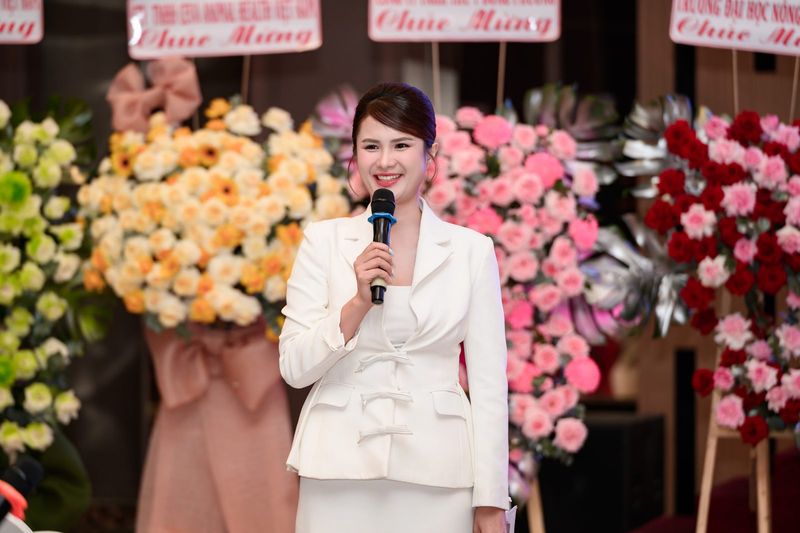 Vũ Hà Trang mặc vest trắng cầm micro bên các lẵng hoa chúc mừng