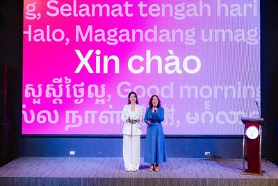 Vũ Hà Trang áo trắng cùng một người dẫn áo xanh trên sân khấu, màn hình lời chào nhiều ngôn ngữ