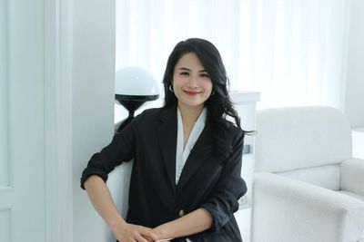 Vũ Hà Trang mặc blazer đen ngồi tựa bên cửa sổ rèm trắng