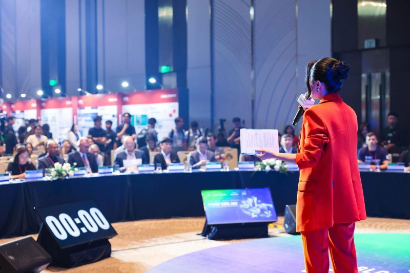 MC áo vest đỏ nhìn từ phía sau dẫn chương trình trước bàn giám khảo Startup Wheel 2025