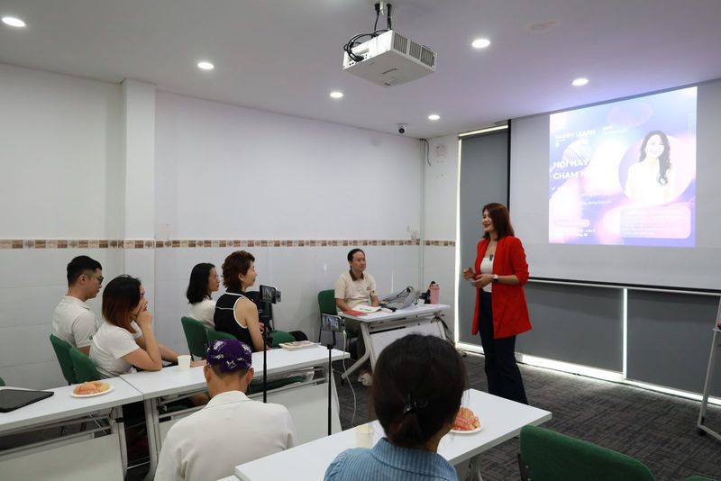 Học viên ngồi quanh bàn trong phòng workshop, Trang đứng cạnh màn chiếu