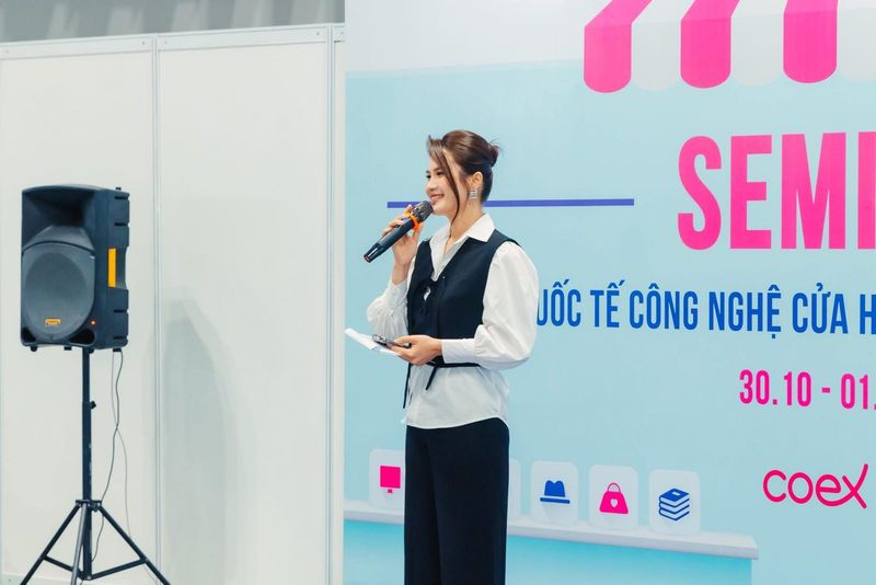 Vũ Hà Trang cầm micro trước backdrop Seminar của triển lãm quốc tế công nghệ cửa hàng