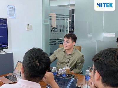 Diễn giả trao đổi với đội kỹ thuật Hitek Software quanh bàn họp, màn hình phía sau hiển thị phiên làm việc dòng lệnh