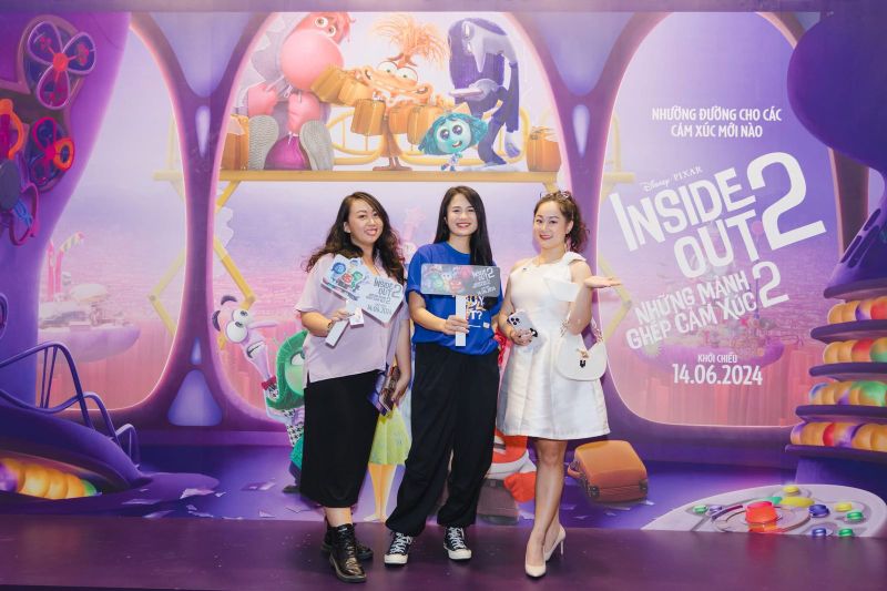 Vũ Hà Trang áo thun xanh đứng giữa hai người trước backdrop Inside Out 2